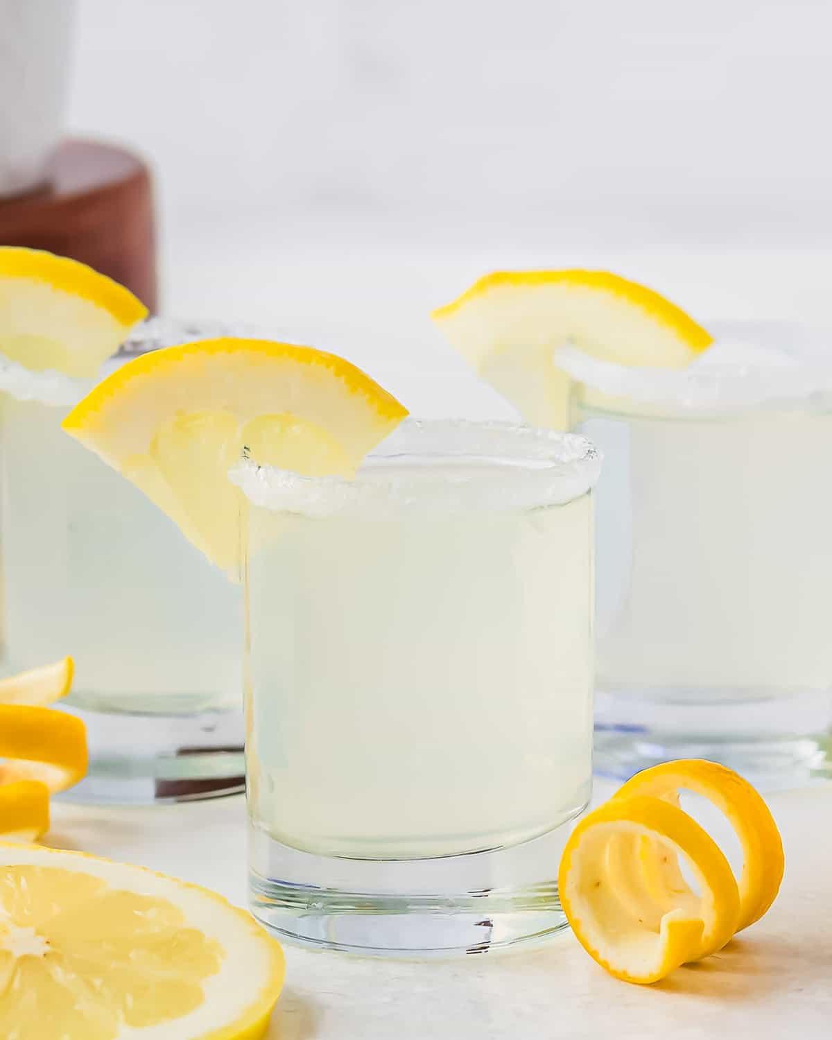 Τρία ποτήρια Lemon Drop Shots, γαρνιρισμένα με φέτες λεμονιού στα χείλη. Τα ποτήρια έχουν ζαχαρωμένα χείλη και είναι τοποθετημένα σε λευκή επιφάνεια, δίπλα σε φέτες και σπείρες λεμονιού. Ένα θολό φόντο με ένα ξύλινο στοιχείο είναι ορατό.