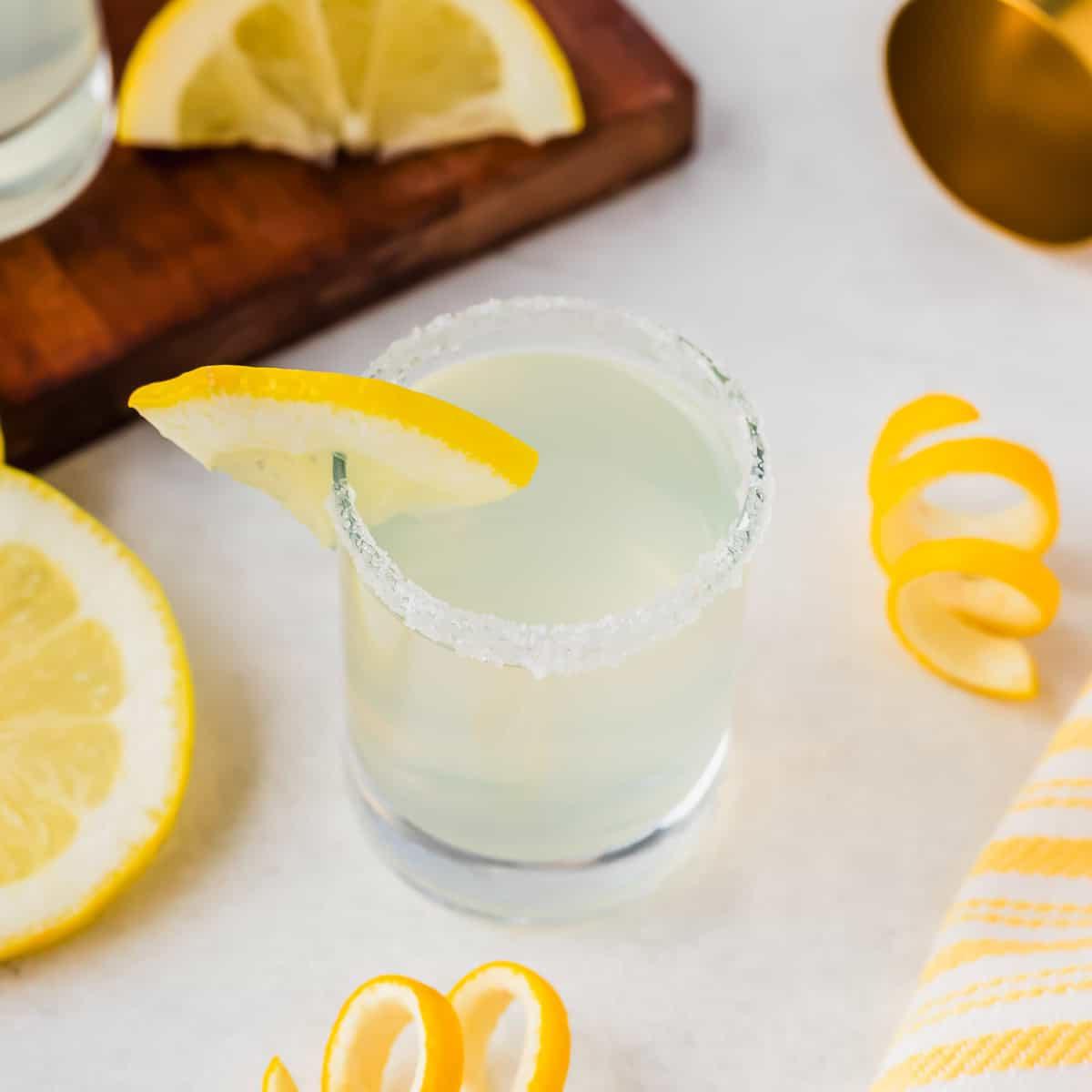 Ένα ποτήρι κοκτέιλ Lemon Drop Shot με ζαχαρωμένο χείλος και γαρνιτούρα φέτας λεμονιού βρίσκεται σε λευκή επιφάνεια. Κοντά βρίσκονται φέτες και φλούδες λεμονιού, ένα κίτρινο ριγέ ύφασμα και ένα ξύλινο σανίδι με περισσότερες φέτες λεμονιού.