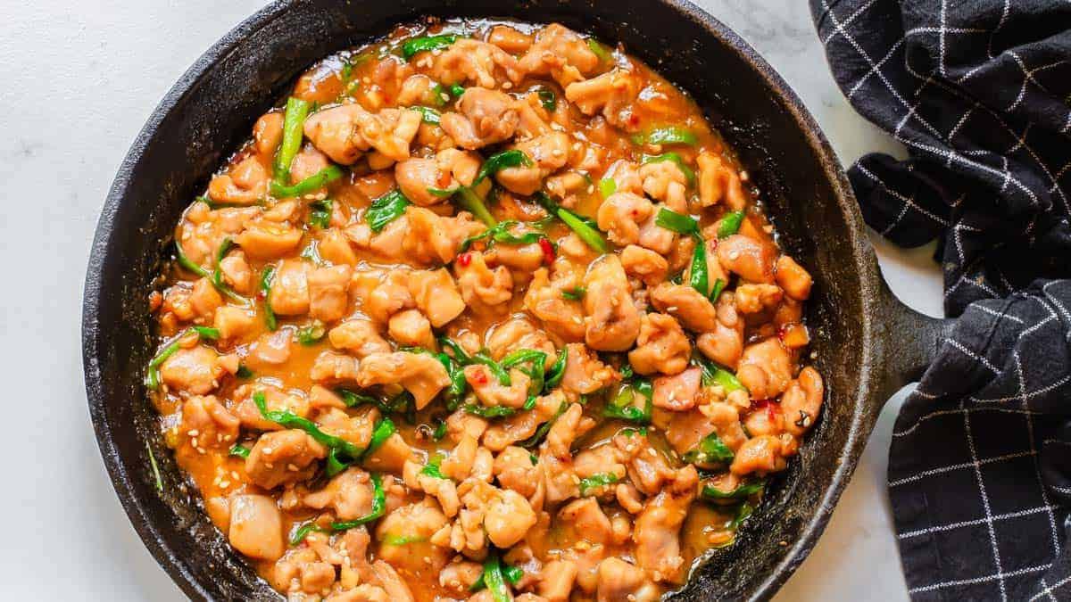 **Wok: 23 Ασιατικά Πιάτα που θα ξεπεράσουν το Takeout** 13 Κινεζικό κοτόπουλο gochujang σε ένα τηγάνι σε ένα τραπέζι με μαύρη πετσέτα κουζίνας τυλιγμένη γύρω από τη λαβή.