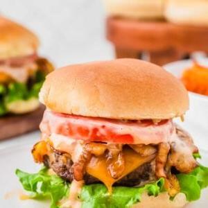 A bison cheeseburger with lettuce, tomato, and sauce on a bun, served on a white plate, with more burgers blurred in the background.