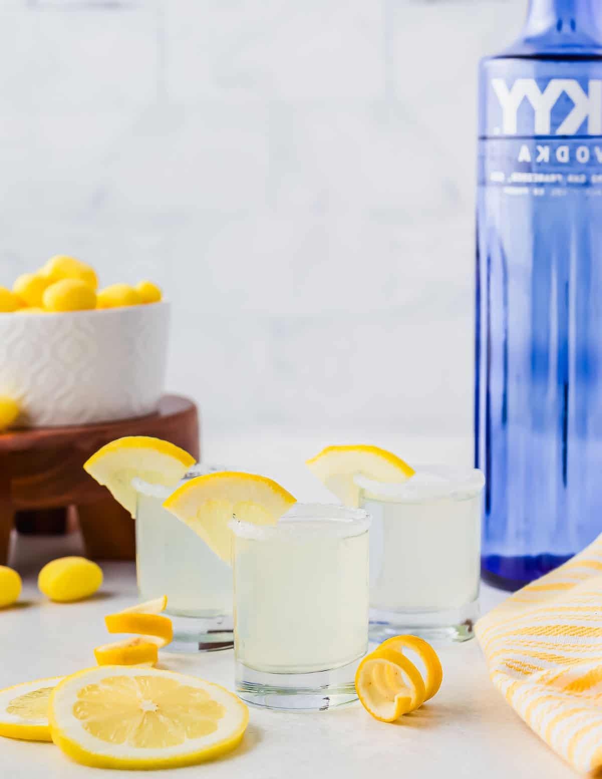 Τρία κοκτέιλ lemon drop σε ποτήρια σφηνάκι, γαρνιρισμένα με φέτες λεμονιού, κάθονται σε λευκή επιφάνεια. Ένα μπλε μπουκάλι βότκας και ένα μπολ με κίτρινες καραμέλες βρίσκονται στο παρασκήνιο. Φέτες λεμονιού είναι σκορπισμένες τριγύρω, μαζί με μια κίτρινη ριγέ πετσέτα.