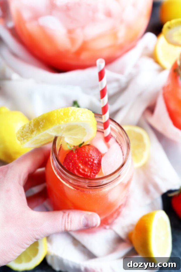 Refreshing Strawberry Vodka Lemonade 6 A hand holding a clear mason jar filled with pink vodka strawberry lemonade, with fresh strawberries and lemon slices in the background.