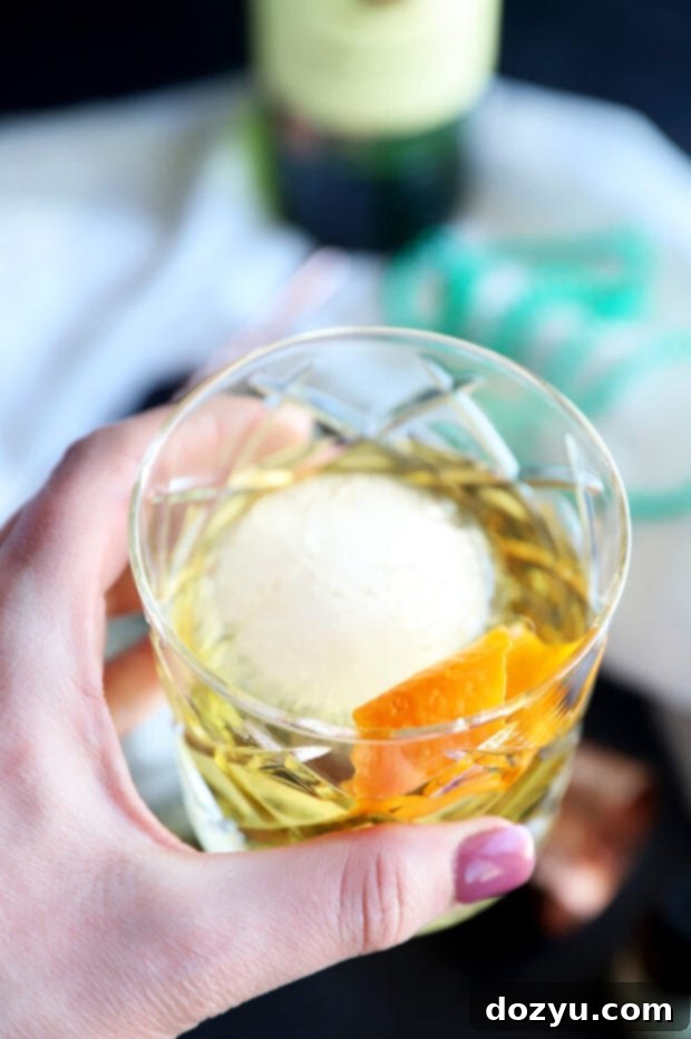 Jameson's Enduring Classic 6 Hand holding a Jameson Old Fashioned cocktail in a rocks glass with a large ice sphere and orange peel garnish, against a dark, moody background.