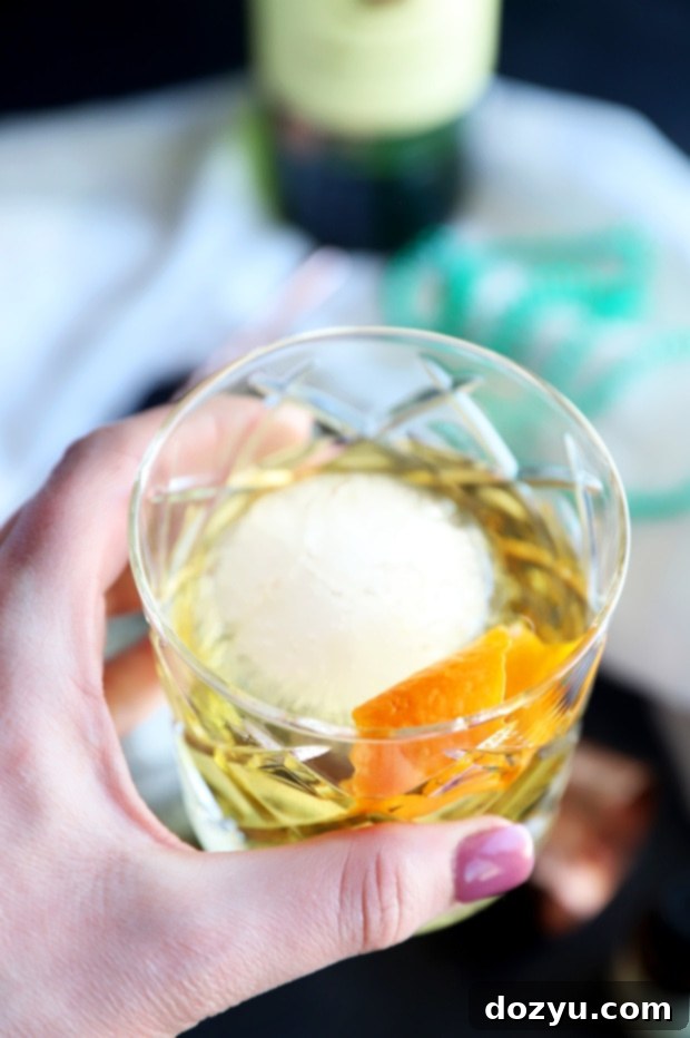 Jameson's Enduring Classic 10 Hand holding a Jameson Old Fashioned cocktail in a rocks glass with a large ice sphere and orange peel garnish, against a dark, moody background.
