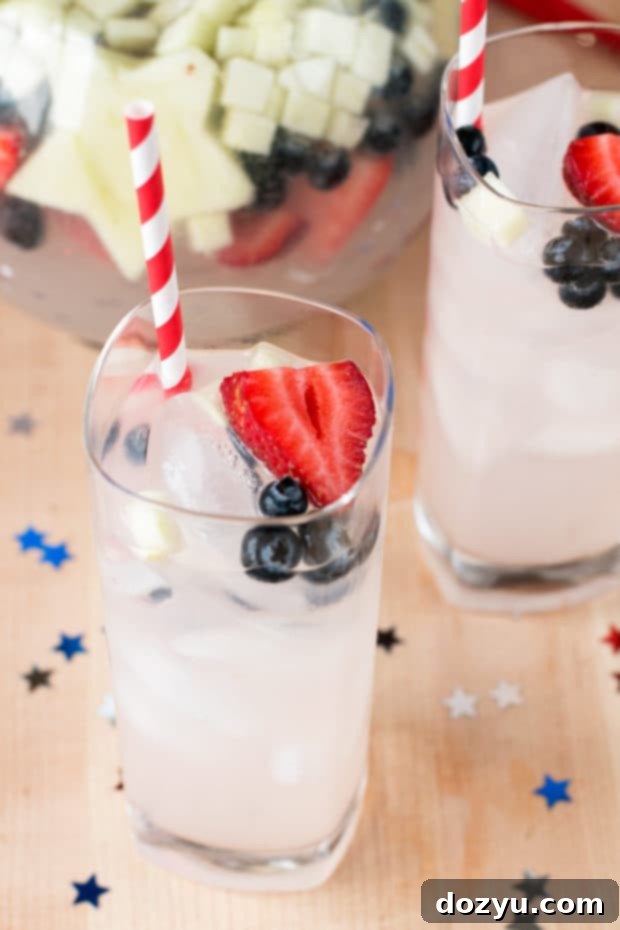 Patriotic Tom Collins Punch overflowing with red strawberries, blue blueberries, and white apple stars in a clear pitcher, perfect for a Fourth of July cocktail party.