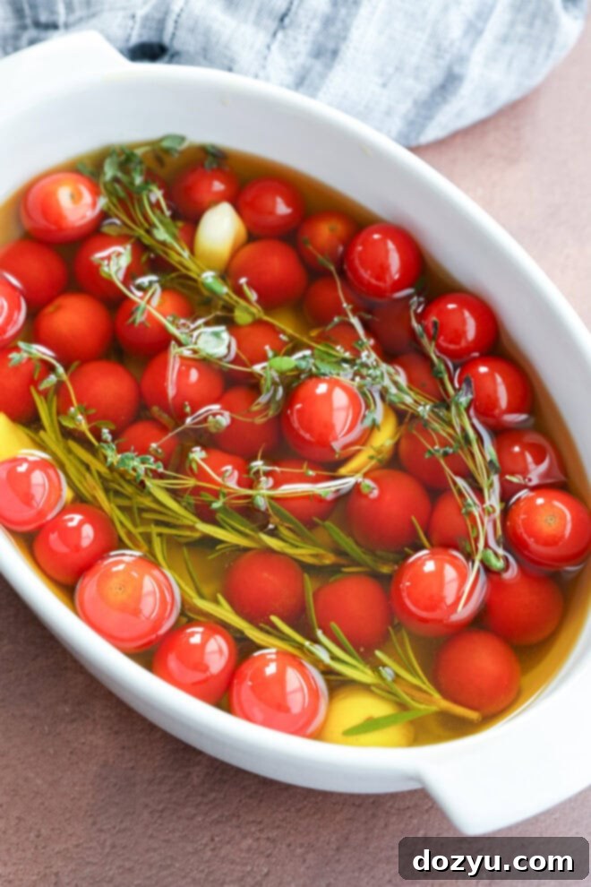 Easy Cherry Tomato Confit 4 A white dish filled with cherry tomato confit, sprigs of fresh rosemary and thyme, and garlic cloves, all submerged in olive oil, before cooking, with a gray cloth in the background.