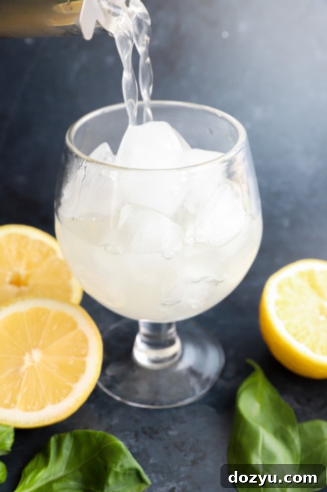 A hand pouring a lemon basil cocktail from a shaker into a glass filled with ice and fresh herbs.