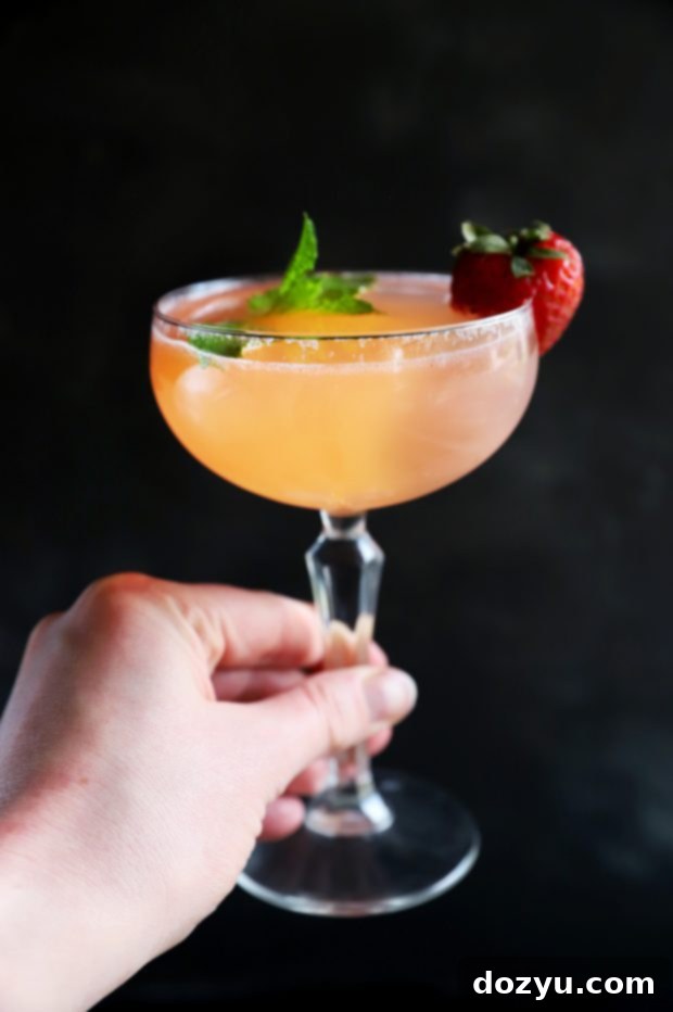 Minted Strawberry Vodka Gimlet 5 A hand holding a perfectly garnished Strawberry Mint Vodka Gimlet in a coupe glass, showcasing its vibrant color and refreshing appeal.