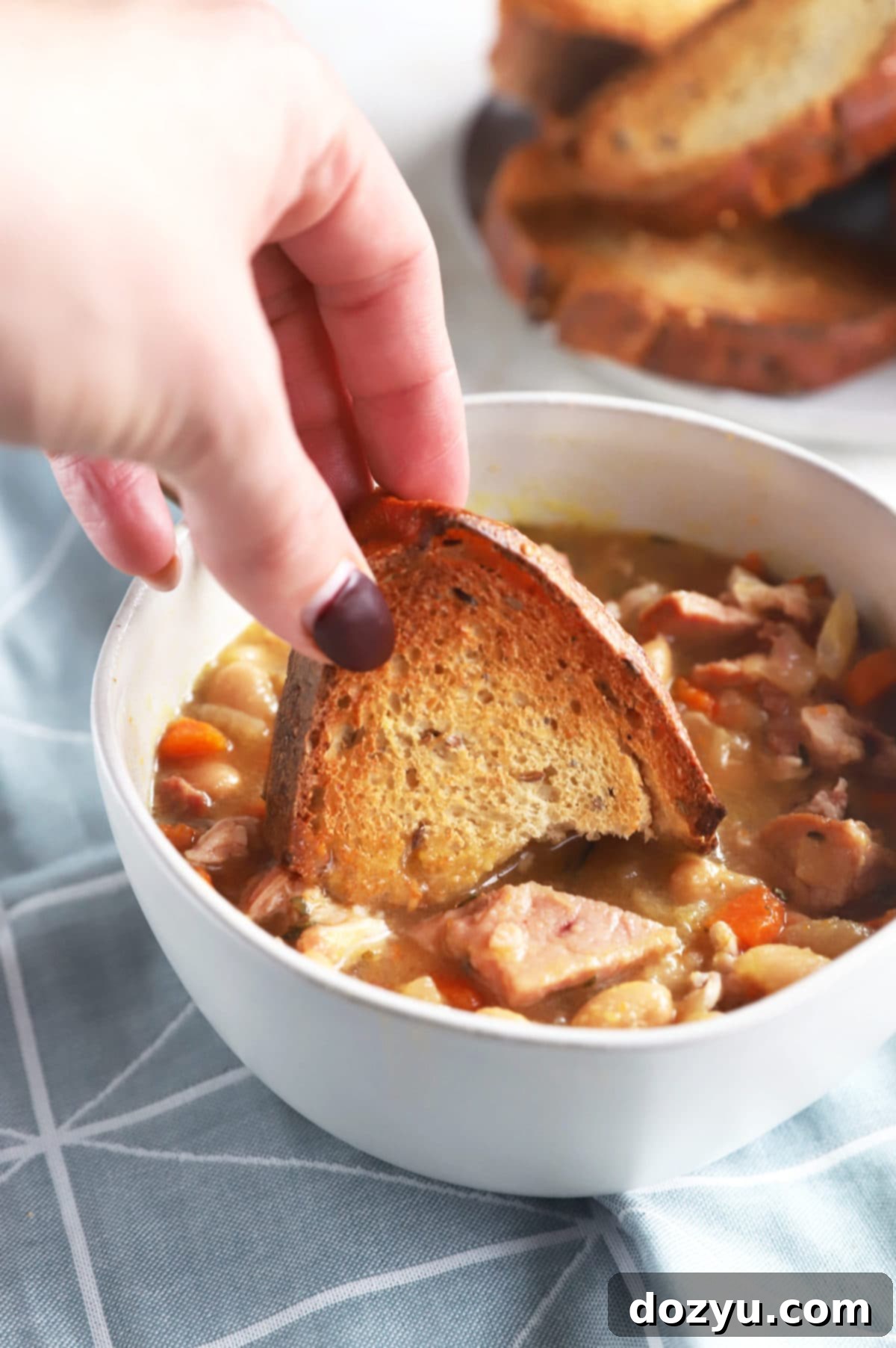 One Pot Ham and Bean Delight 5 A hand dipping a piece of crusty bread into a bowl of Instant Pot Ham and Bean Soup, highlighting the rich texture.