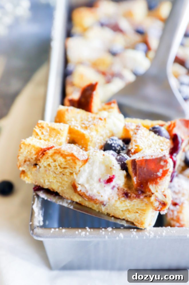 Blueberry & Cream Cheese French Toast Bake 2 Spatula with a golden slice of Blueberry Cream Cheese French Toast Casserole, topped with powdered sugar and a drizzle of strawberry coulis.