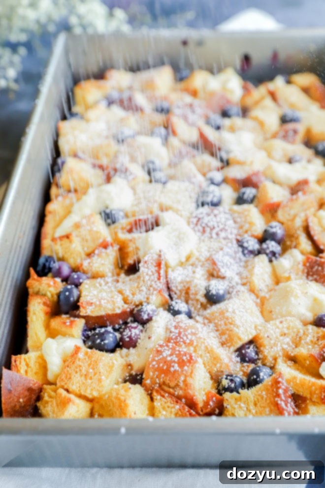 Blueberry & Cream Cheese French Toast Bake 6 A hand sprinkling powdered sugar over a freshly baked Blueberry Cream Cheese French Toast Casserole in a pan.