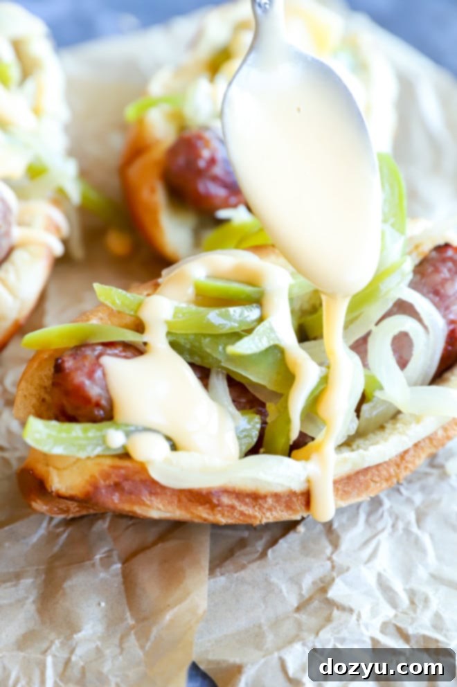 Philly Cheesesteak Beer Brats 8 Hand drizzling creamy cheese sauce over a fully assembled grilled bratwurst, topped with beer-braised vegetables, in a bun.