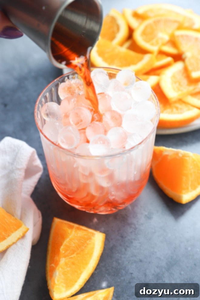 Aperol Fizz 5 Hand pouring vibrant Aperol into a cocktail glass filled with ice, with fresh orange slices in the background, preparing an Aperol Soda.