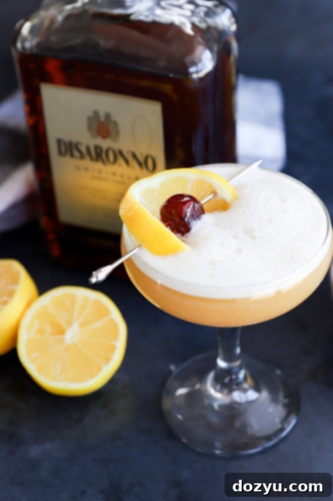 The Disaronno Sour 2 Disaronno sour cocktail in a coupe glass with lemon and cherry garnish and disaronno bottle in the background