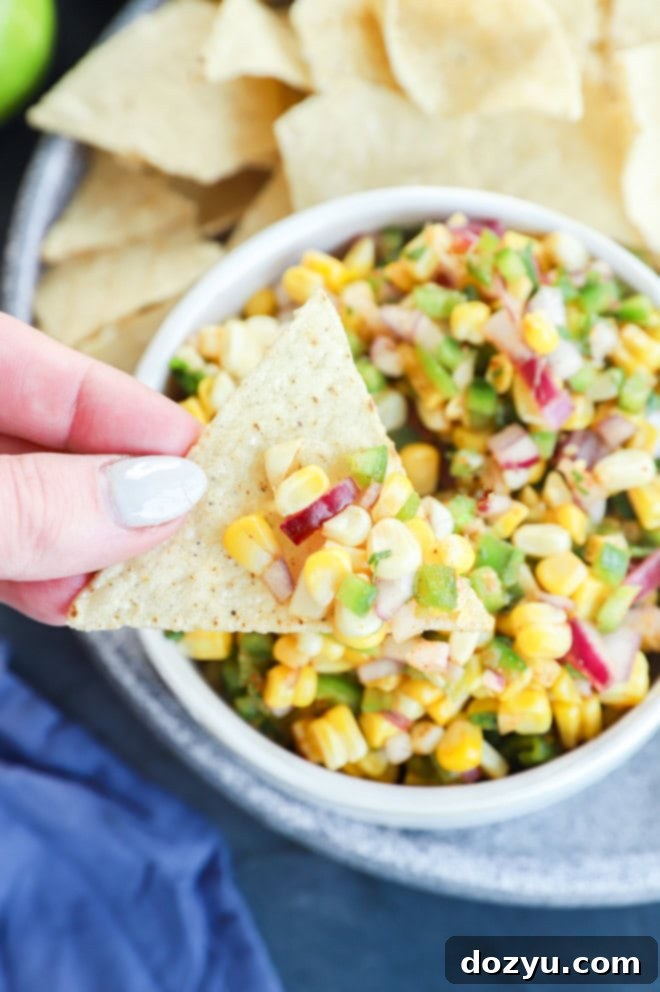 Smoky Roasted Corn Salsa 8 Hand holding a chip with roasted chili corn salsa in a bowl and tortilla chips on a plate