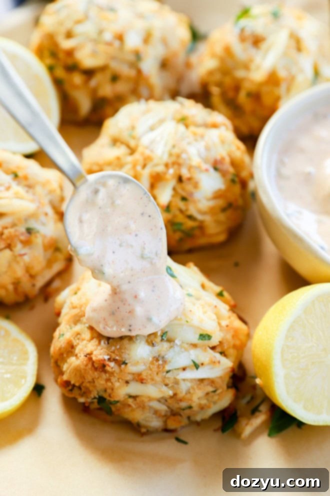 Effortless Crab Cake Sauce 6 spooning creamy, tangy crab cake sauce onto a golden baked seafood appetizer, with fresh lemon wedges for garnish.
