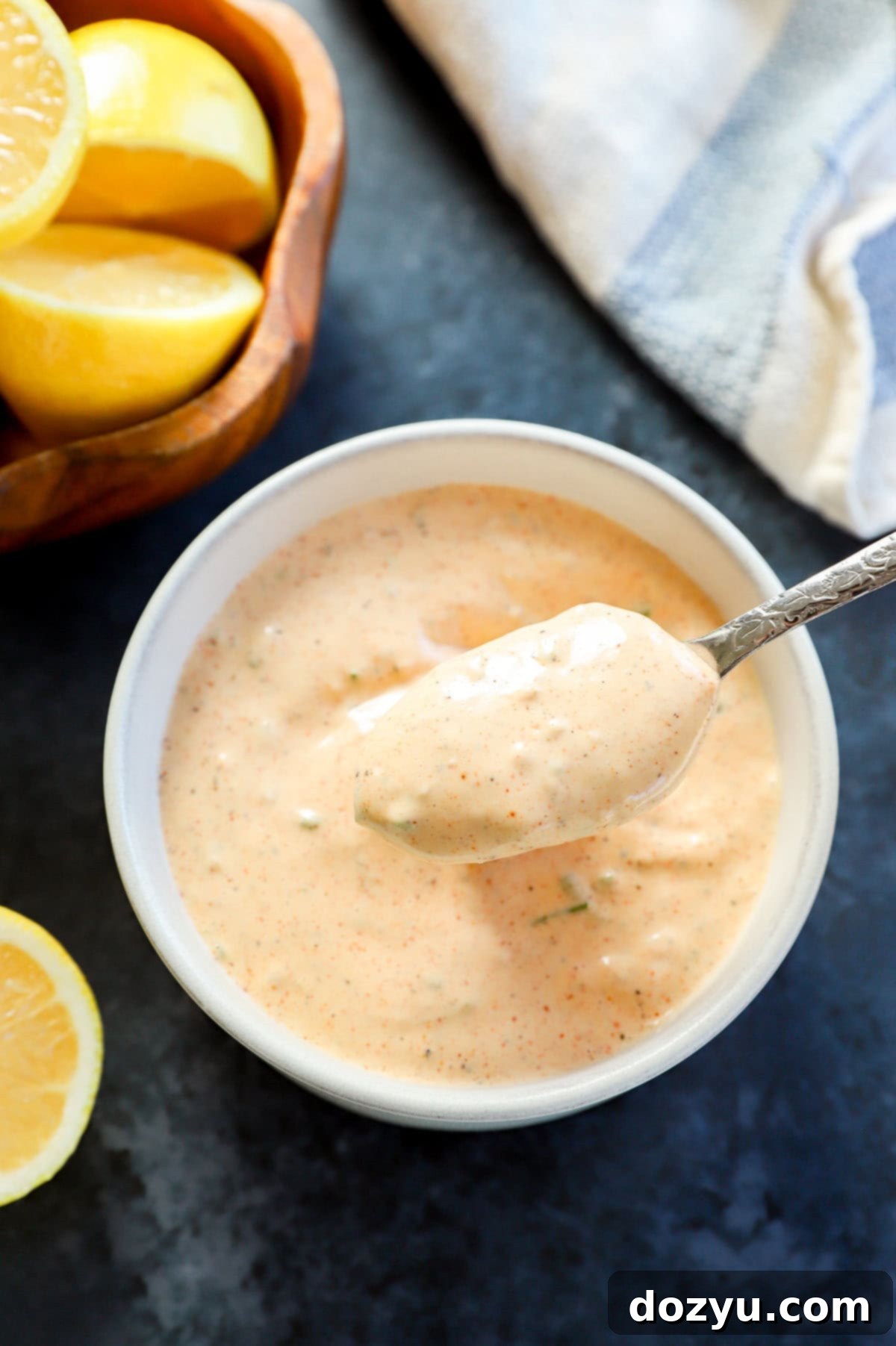 Effortless Crab Cake Sauce 8 spoon lifting out creamy remoulade sauce for crabs cakes from a bowl, with a bowl of lemon halves nearby.