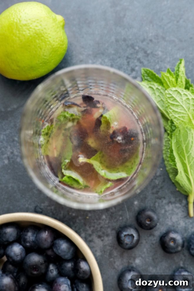 Blueberry Mojito Magic 4 Close-up of a cocktail glass with muddled fresh blueberries, lime slices, raw sugar, and mint leaves at the bottom, ready for rum and soda.