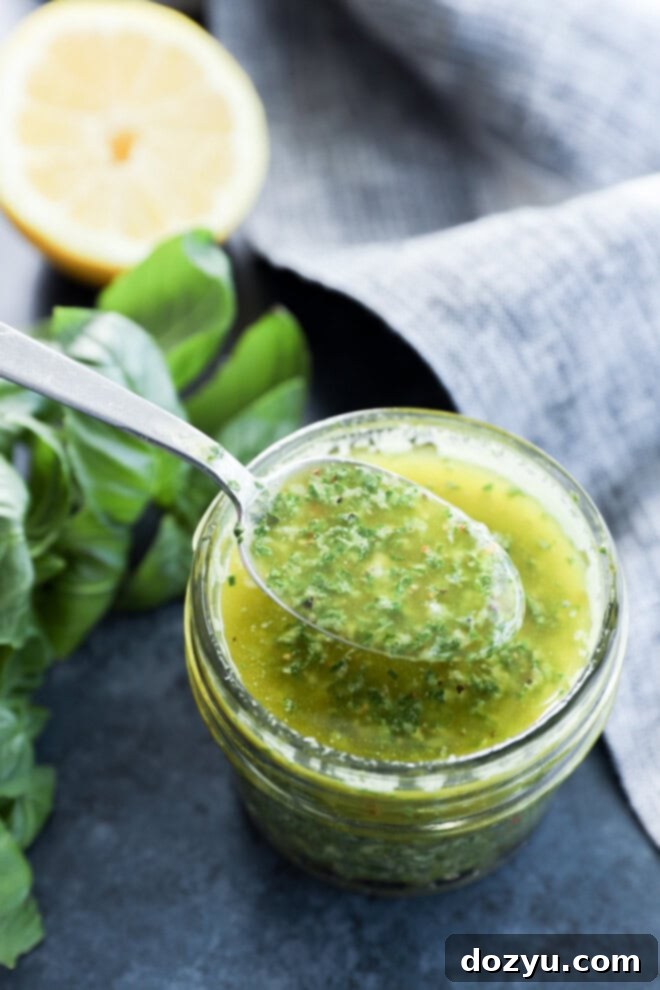 The Magic of Basil Oil 4 A spoon is scooping vibrant green basil oil from a mason jar, with fresh basil sprigs and a lemon half blurred in the background, set on a rustic surface.