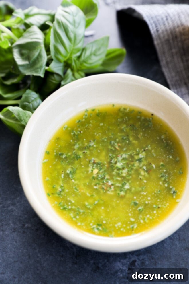 The Magic of Basil Oil 5 A vibrant green basil oil in a small white bowl, garnished with a fresh sprig of basil and red pepper flakes, against a grey linen background with additional herbs.