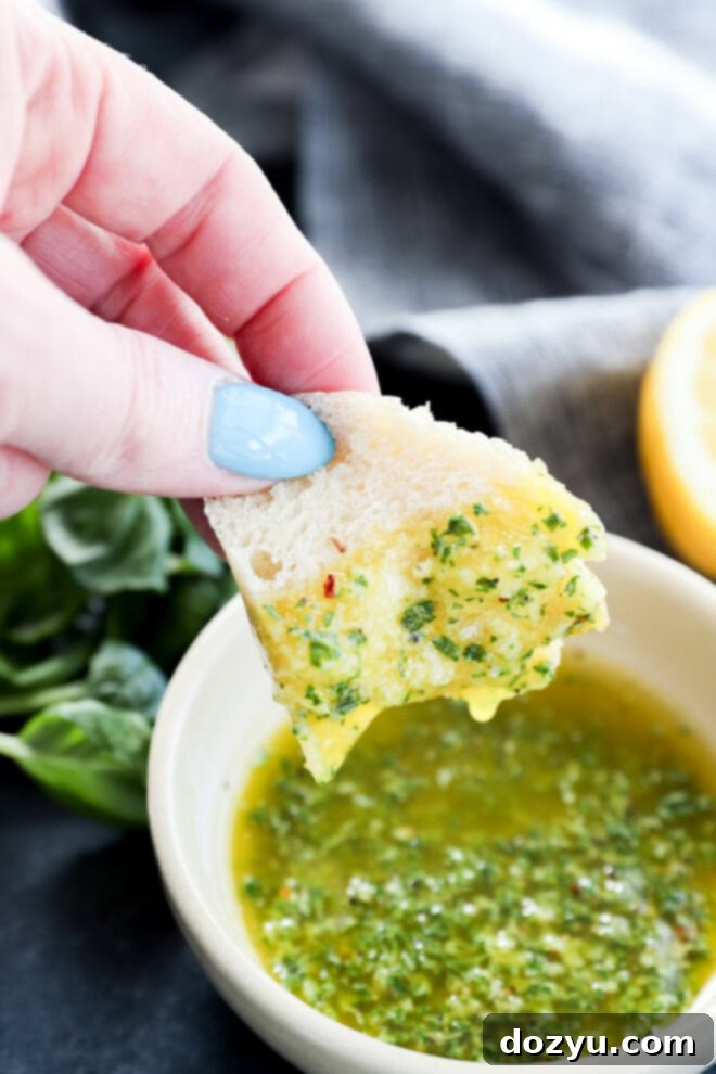The Magic of Basil Oil 6 A hand dipping a piece of rustic bread into a bowl of fresh basil oil, with fresh basil and lemon in the background.