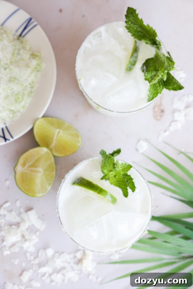 Coconut Mojito 8 An inviting overhead view captures two glasses brimming with a white, frothy Coconut Mojito, each beautifully garnished with fresh mint and delicate lime slices. Adjacent on a clean, light surface, are more vibrant lime wedges, a plate featuring a subtly powdery lime-sugar mixture, and a lush green palm leaf, all contributing to a captivating tropical drink presentation.