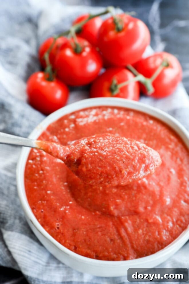 Ultimate Pizza Sauce 2 A spoonful of thick, Best Pizza Sauce is held above a bowl filled with more sauce, with fresh vine tomatoes in the background on a striped cloth.