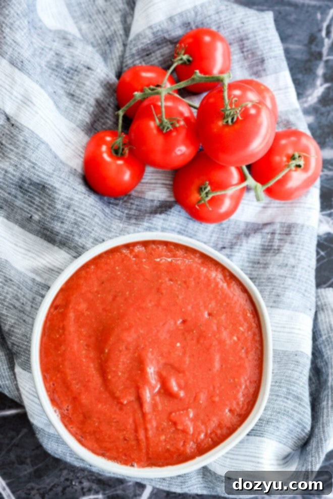 Ultimate Pizza Sauce 6 A cluster of fresh tomatoes on the vine sits next to a bowl of smooth tomato sauce arranged on a striped gray and white cloth atop a dark marble surface.
