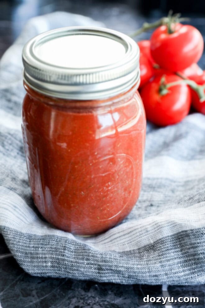 Ultimate Pizza Sauce 7 A glass mason jar filled with red tomato sauce, known as the best pizza sauce, sits on a striped cloth, with fresh tomatoes on the vine in the background.
