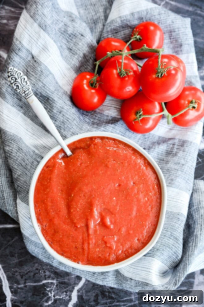 Ultimate Pizza Sauce 9 A bowl of smooth red tomato sauce, perfect as the best pizza sauce, with a spoon rests on a striped cloth beside fresh tomatoes on the vine, all set on a dark marble surface.