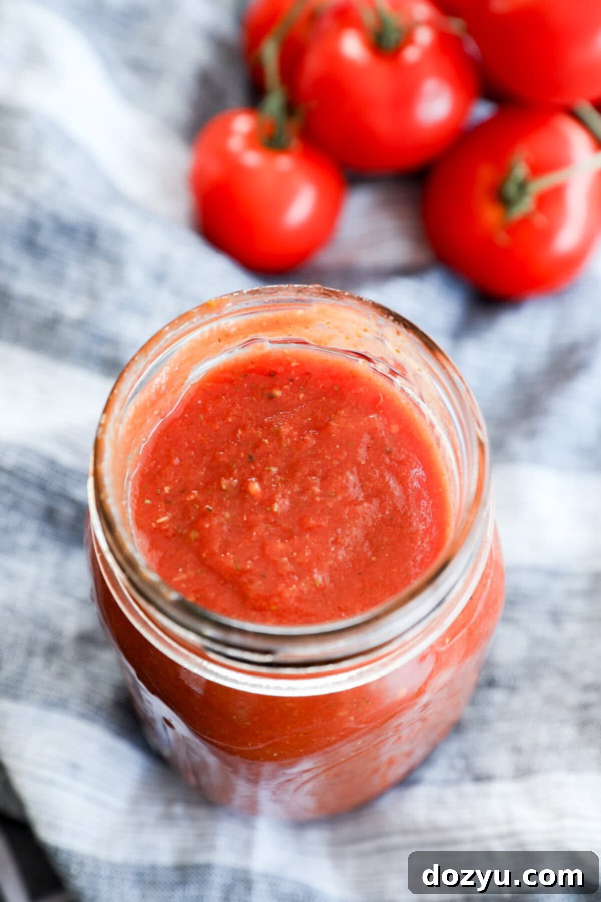 Ultimate Pizza Sauce 10 A glass jar filled with red tomato sauce, perfect as the best pizza sauce, sits on a striped cloth, with several fresh tomatoes on the vine in the background.