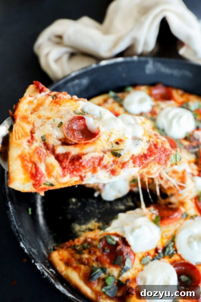 Sizzling Skillet Pepperoni Pie 8 A slice of spicy pizza with melted cheese is being lifted from a pan, topped with dollops of ricotta and fresh basil. A beige cloth is in the background.