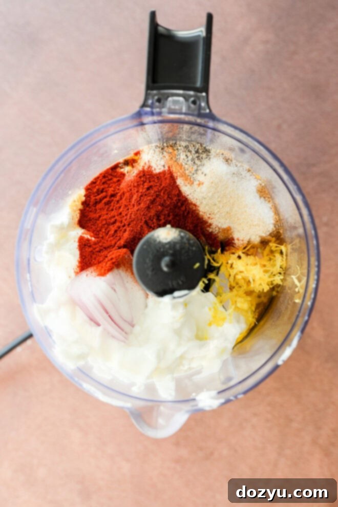 Perfect Greek Yogurt Chicken Marinade 4 A food processor bowl filled with a creamy mix of Greek yogurt, sweet paprika, garlic powder, lemon zest, roughly chopped shallot, and olive oil, viewed from directly above on a light brown textured surface, showing the fresh ingredients pre-blending.