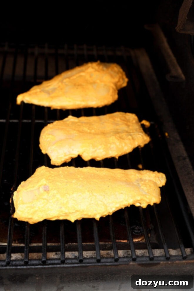 Perfect Greek Yogurt Chicken Marinade 7 Three pieces of chicken, richly coated in a vibrant yellow Greek yogurt marinade, are actively cooking on a hot grill, showcasing distinct, appetizing grill marks on the grates beneath them, promising a smoky, flavorful meal.