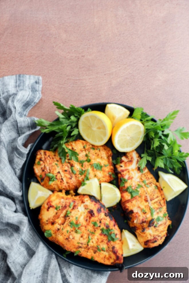 Perfect Greek Yogurt Chicken Marinade 8 A black plate beautifully presents three grilled chicken breasts, generously garnished with fresh chopped herbs, artfully surrounded by vibrant lemon wedges and fresh parsley sprigs, set on a rustic textured brown surface next to a soft gray cloth napkin.