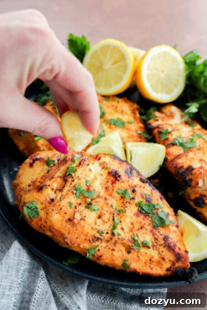 Perfect Greek Yogurt Chicken Marinade 9 A hand gently squeezes a lemon wedge over perfectly grilled Greek yogurt chicken breasts, garnished with fresh chopped herbs, with more lemon wedges and fresh parsley in the background on a sleek black plate.