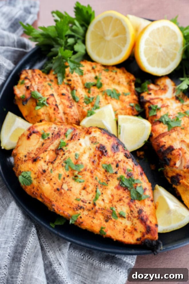 Perfect Greek Yogurt Chicken Marinade 10 Grilled, seasoned Greek yogurt chicken breasts, garnished with fresh chopped herbs, served elegantly on a black plate with vibrant lemon wedges and fresh parsley sprigs, positioned next to a stylish gray and white striped cloth for a complete and appealing presentation.