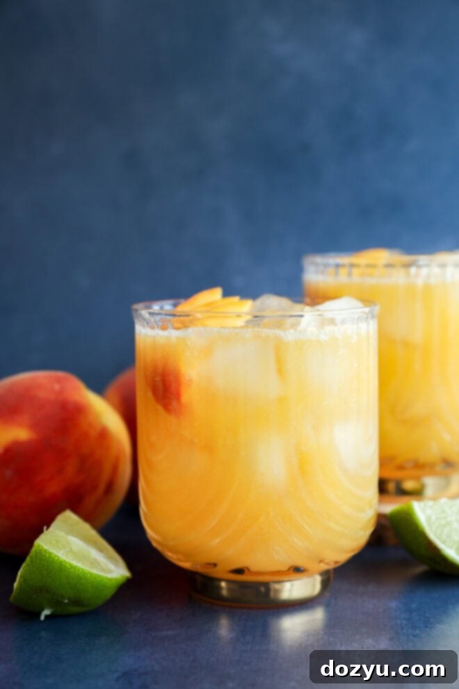 Two glasses of vibrant peach margarita, garnished with fresh peach slices, sit on a dark surface. Nearby are whole ripe peaches and a halved lime, all against a softly blurred blue background, inviting a refreshing sip.