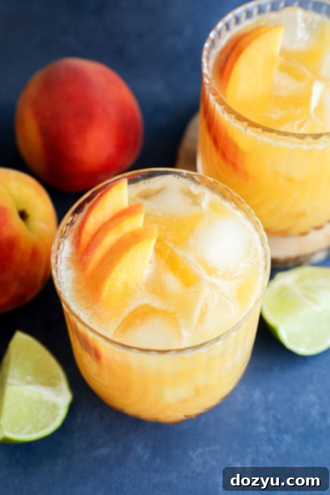 Two glasses of refreshing iced peach margarita cocktails, elegantly garnished with fresh peach slices and a lime wedge, are beautifully arranged on a blue surface. Fresh whole peaches and lime wedges provide a vibrant backdrop, inviting a taste of summer.