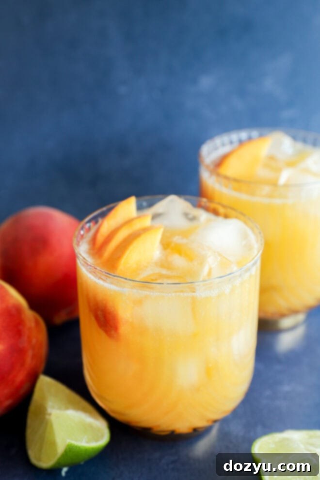 A single glass of iced peach margarita, beautifully garnished with fresh peach slices, rests on a blue surface. In the background, another similar glass, along with whole peaches and lime wedges, creates a refreshing and inviting scene.