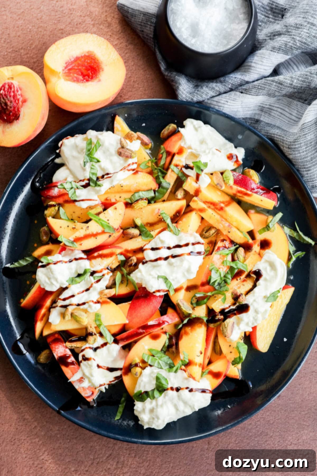 Velvety Burrata with Sun-Ripened Peaches 9 A black plate with sliced peaches, strips of red bell pepper, dollops of creamy cheese, chopped basil, pistachios, and a drizzle of balsamic glaze, next to a halved peach and a small bowl of flaky salt.
