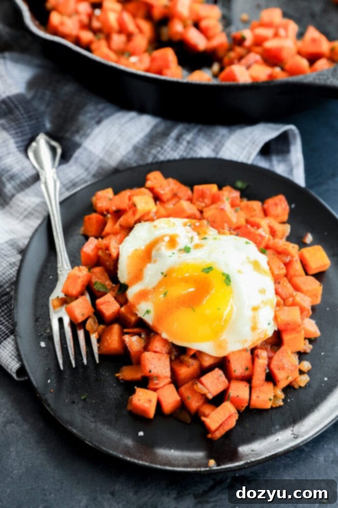 Sweet Potato Sunrise 7 A black plate with diced sweet potatoes topped with a sunny-side-up egg and herbs, next to a fork on a plaid cloth, with a skillet of more sweet potatoes in the background.