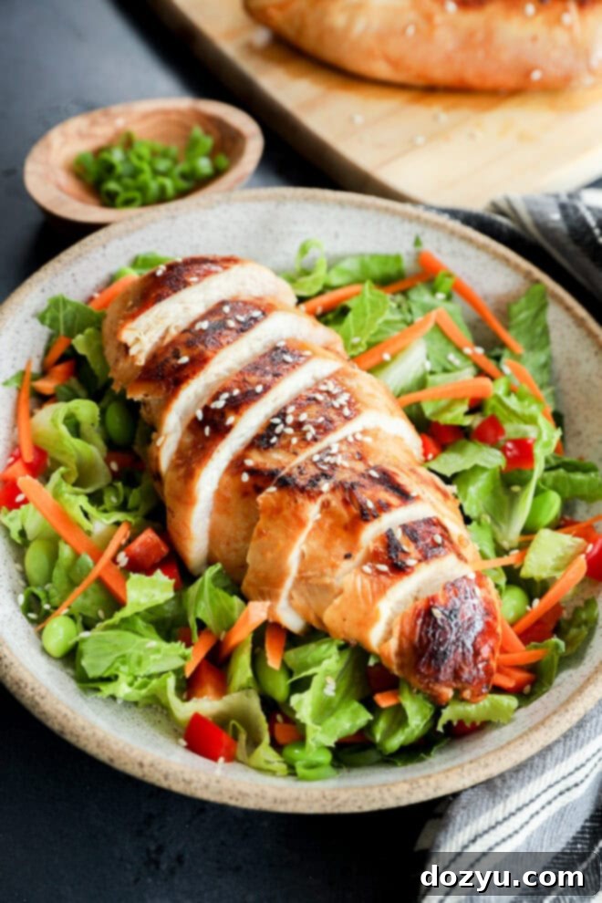 Savory Asian Chicken Marinade 7 A plate with sliced grilled chicken breast, marinated in an Asian Chicken Marinade, served over mixed greens, shredded carrots, red bell peppers, and edamame, garnished with sesame seeds. A wooden bowl with chopped green onions sits in the background.
