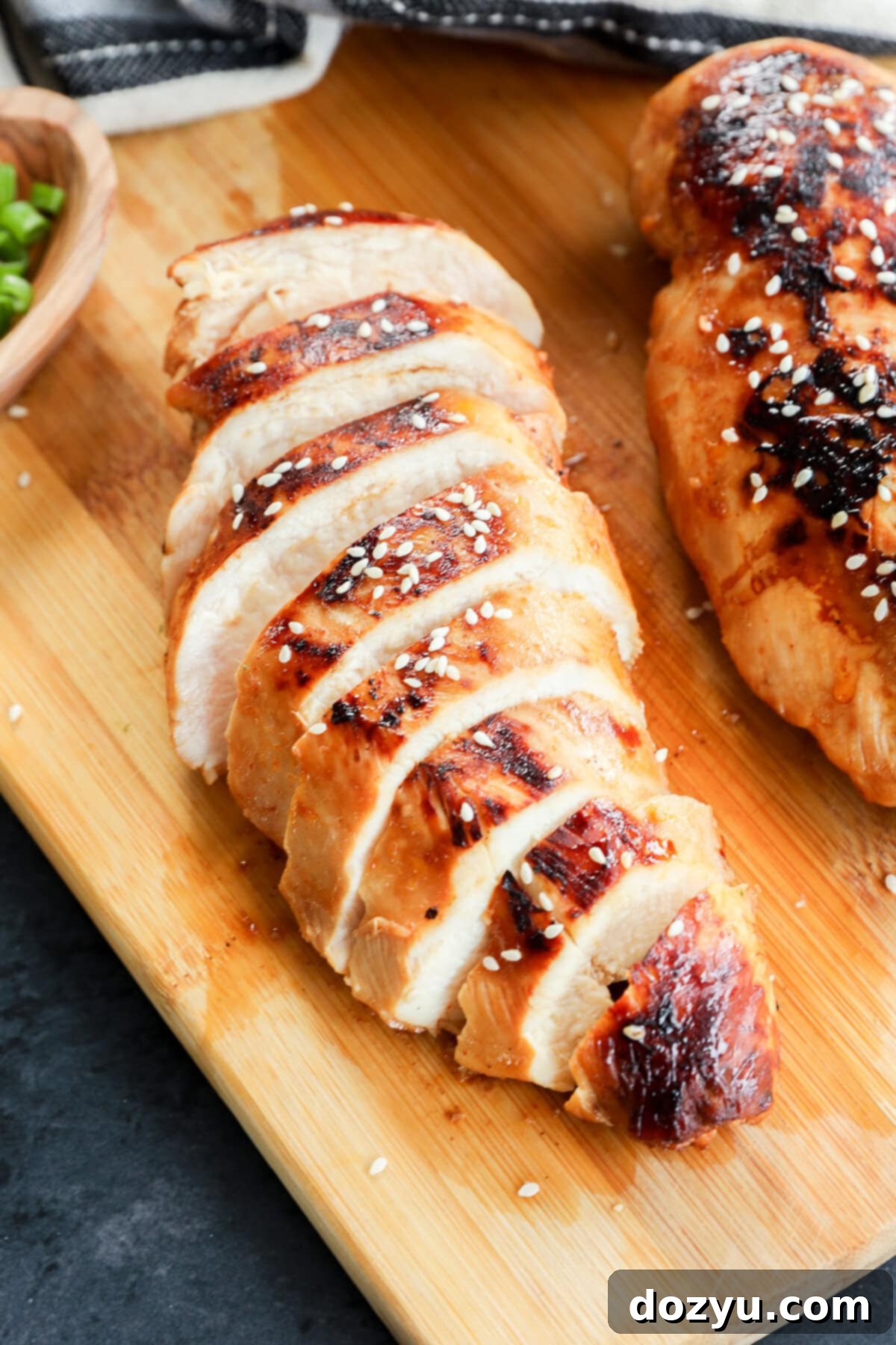 Sliced grilled chicken breast, topped with sesame seeds, rests on a wooden cutting board, with part of another chicken breast and a small bowl of green onions nearby.