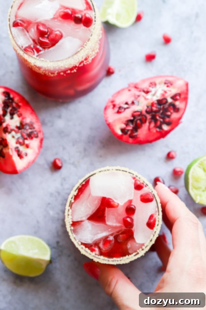 A hand holding a garnished pomegranate margarita in a clear glass, with a festive backdrop.