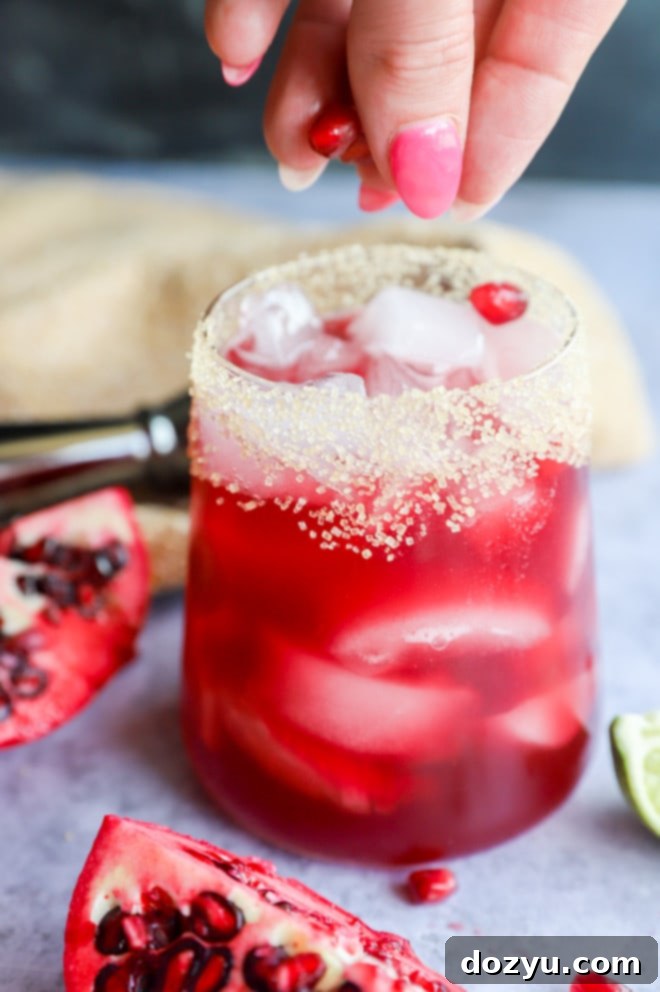 A hand garnishing a pomegranate margarita with fresh pomegranate arils and a lime wedge.