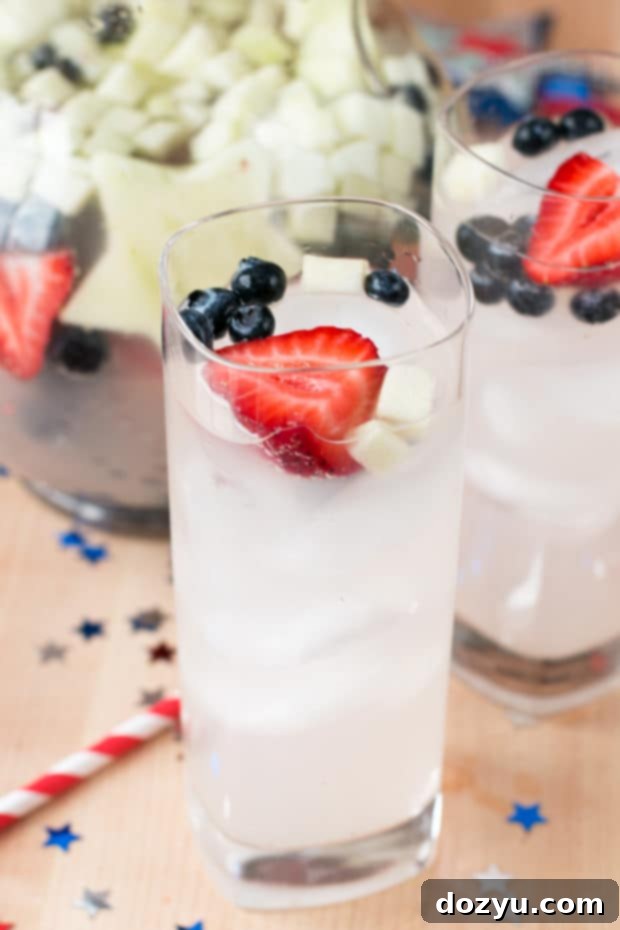 A glass of Patriotic Tom Collins Punch with ice and garnishes, with a pitcher in the background
