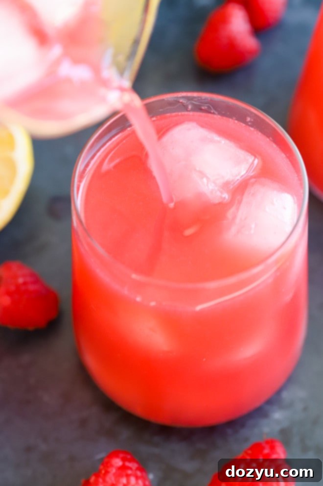 A hand pouring vibrant raspberry lemonade from a pitcher into a glass filled with ice and fresh lemon slices.