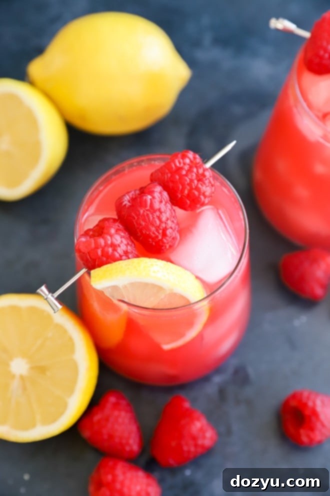 Close-up of a glass filled with raspberry lemonade, ice, fresh raspberries, and a lemon slice, highlighting the vibrant color and refreshing appeal.