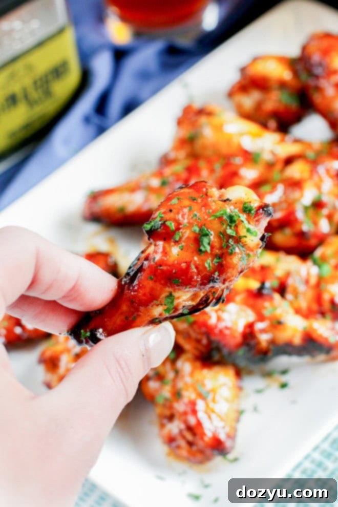 Close-up of a hand holding a perfectly grilled chicken wing, coated in BBQ sauce with spices sprinkled in the background.